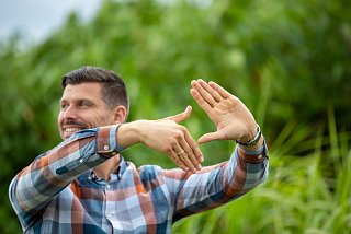 Man stands in nature and forms a rhombus with his hands and smiles to the side