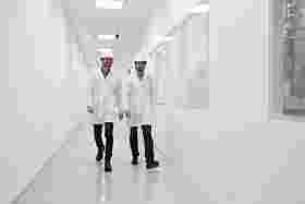 Two men in protective clothing walk down a corridor smiling