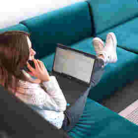 Frau sitzt auf einem Sofa, telefoniert und arbeitet am Laptop
